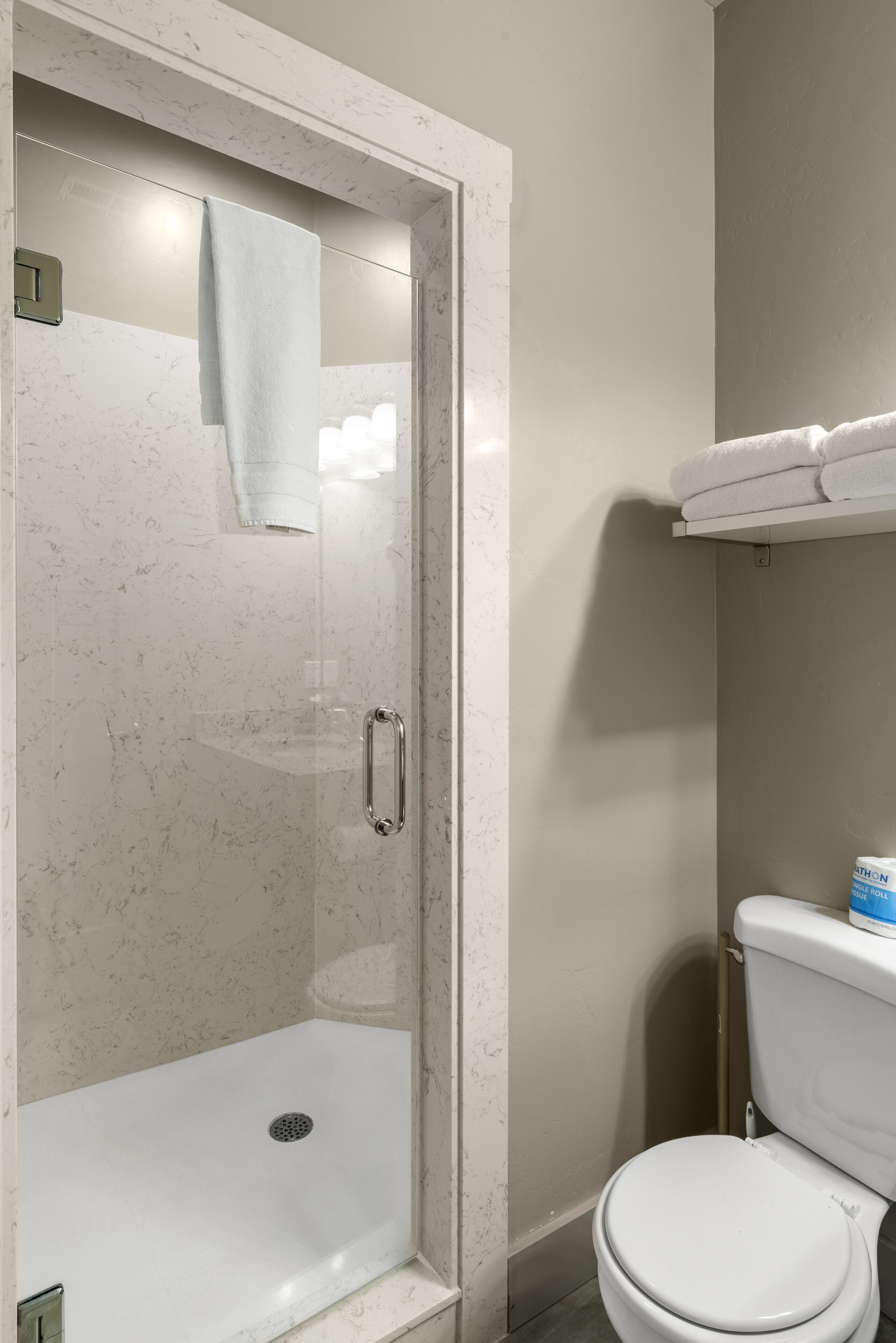 A bathroom with a glass-enclosed shower, a toilet, and a shelf with a rolled-up towel.