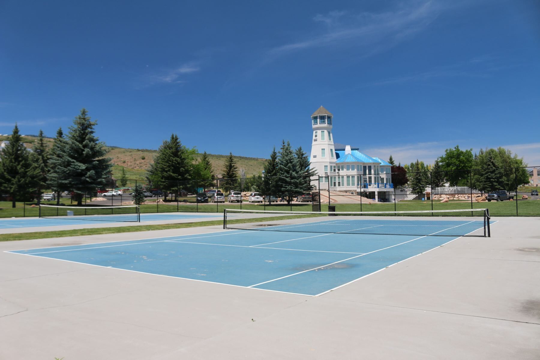An outdoor tennis court with a net, surrounded by a fence, and a building with a tower in the background.