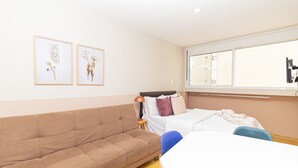 A room with a brown sofa, a bed with white bedding, a small table with a lamp, and two framed floral artworks on the wall. - Vossa Bossa Bela Cintra Home (São Paulo)