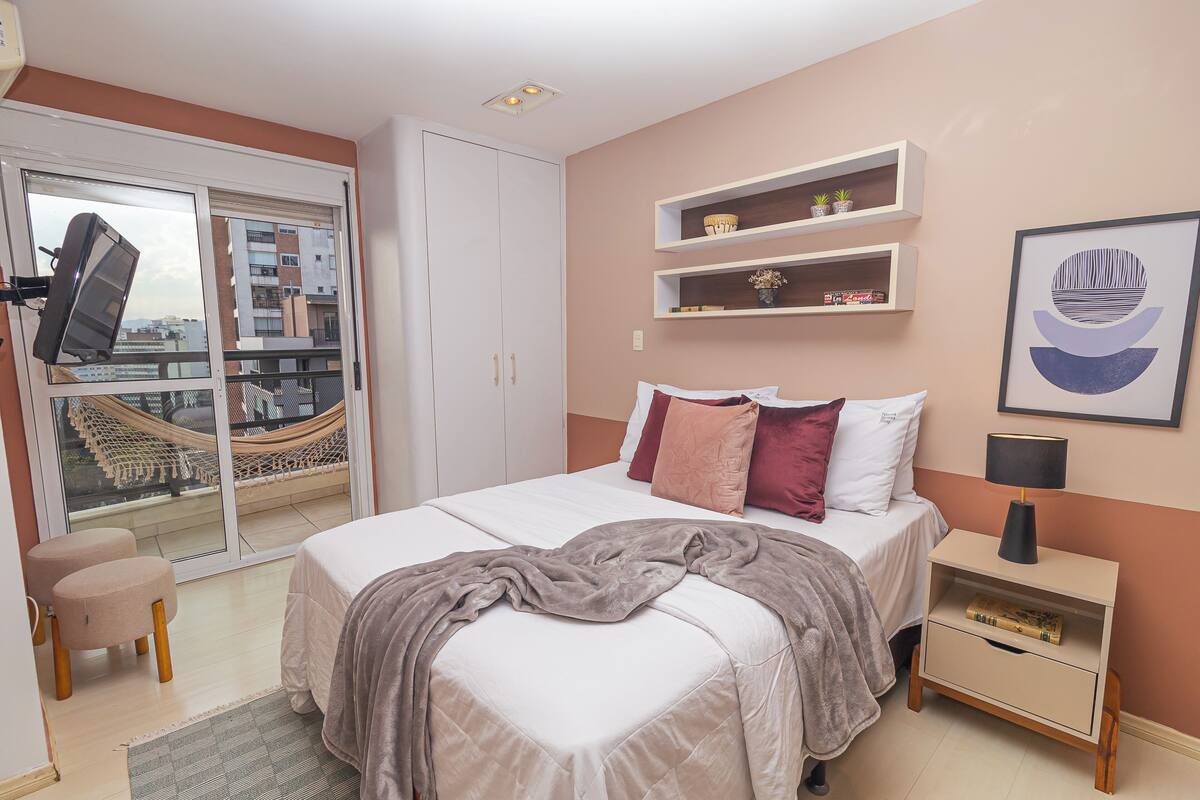 Une chambre à coucher avec un lit, des tables de chevet, une fenêtre donnant sur des immeubles et un hamac à l’extérieur.