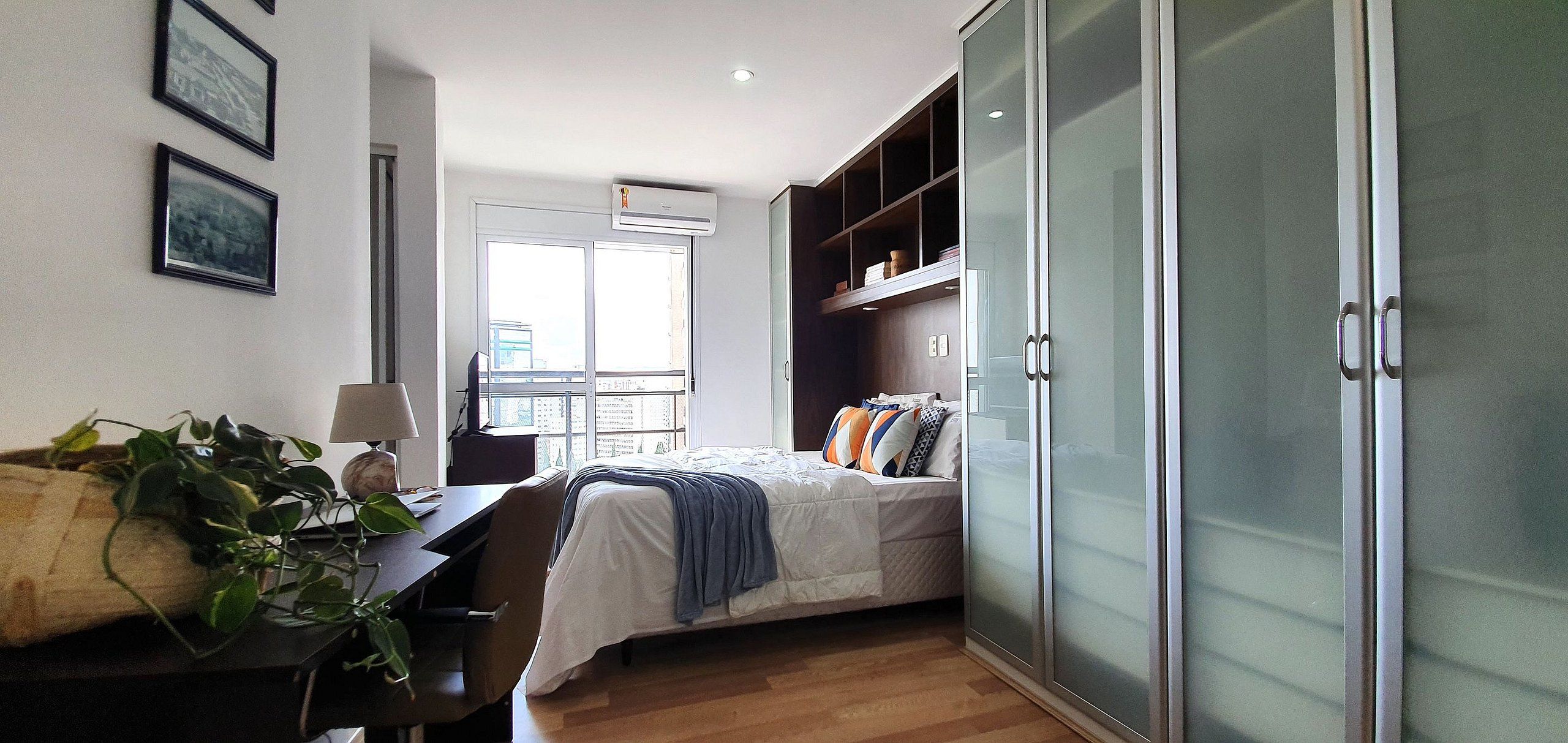 Un dormitorio con cama, escritorio, silla, planta, armario y ventana.