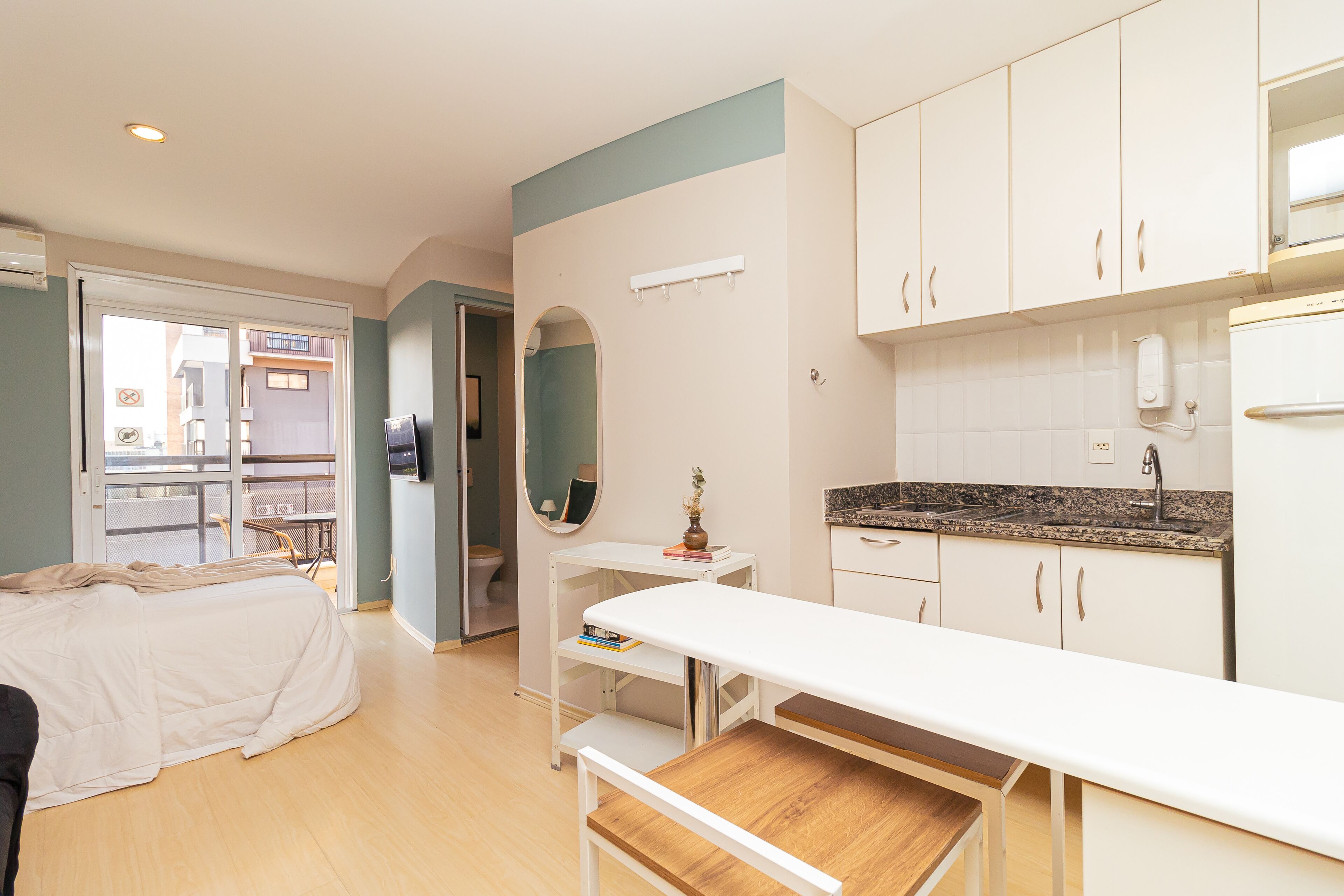 A modern hotel room with a bed, a desk, a small table, a kitchenette, and a bathroom with a mirror and toilet.