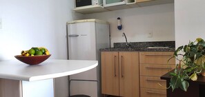 Een kleine keuken met een witte koelkast, een magnetron en een spoelbak. Op een witte tafel staat een fruitschaal en op het aanrecht een bloempot.