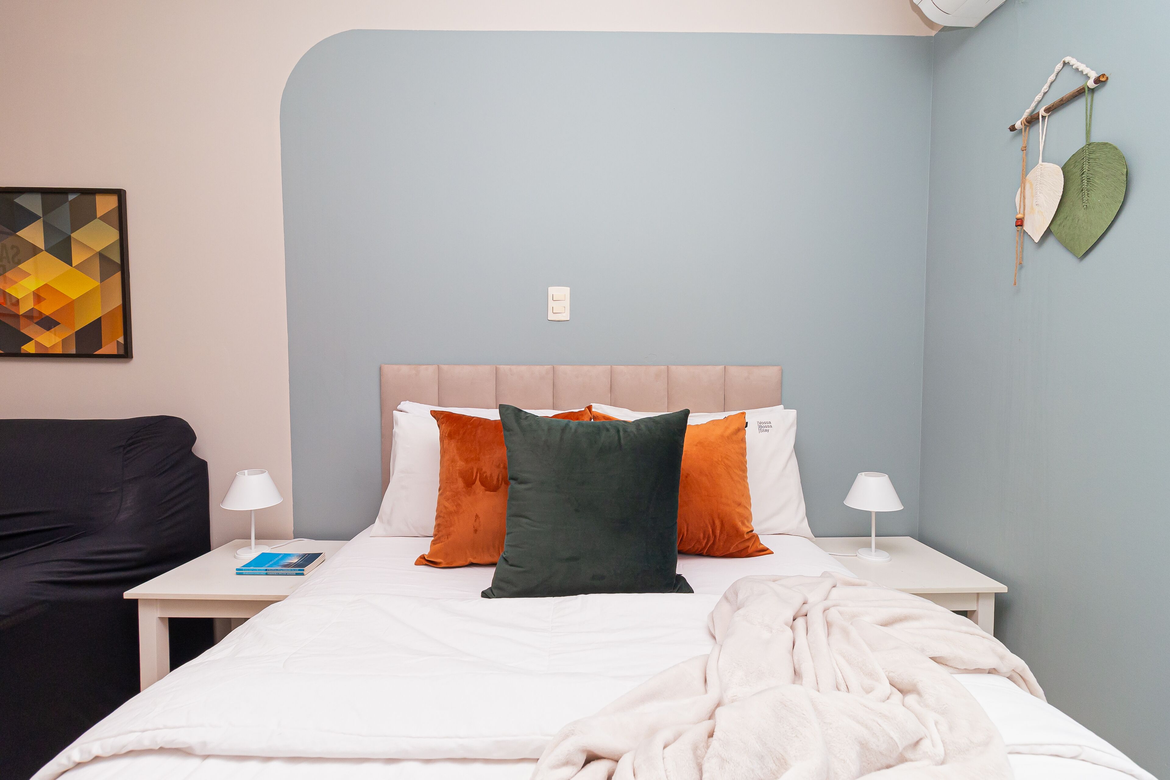A neatly made bed with a white comforter and pillows, a bedside table with a lamp, and a wall-mounted decorative item.