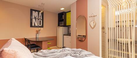 A bedroom with a bed, a desk, a chair, a refrigerator, a mirror, and a macramé curtain.