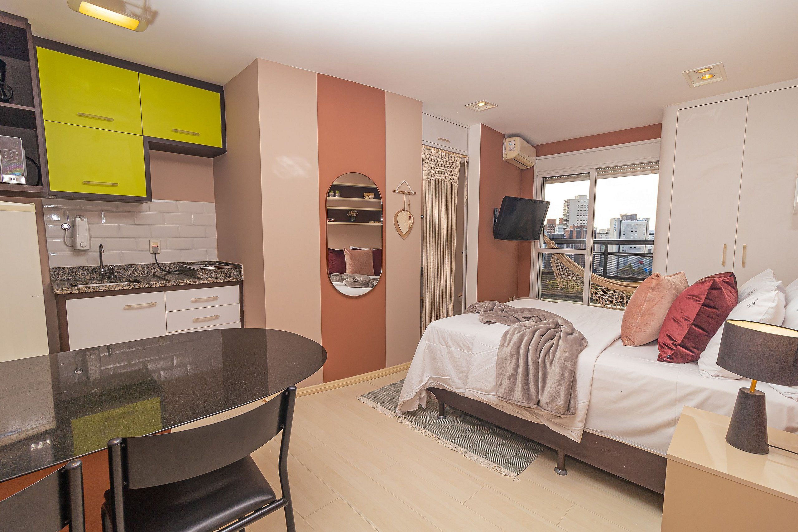 A compact hotel room with a kitchenette, a bed with multiple pillows, a TV, and a window with a city view.