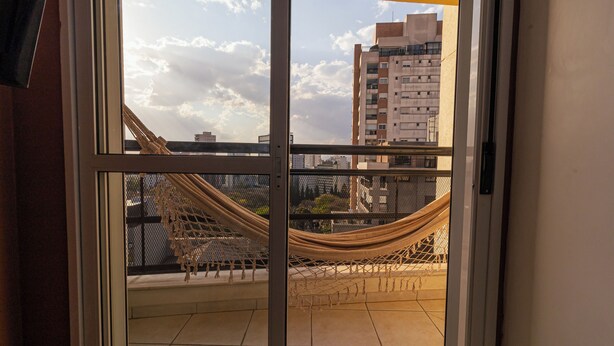 A balcony with a hammock and a view of a cityscape through a glass door. - Vossa Bossa Bela Cintra Home (São Paulo)