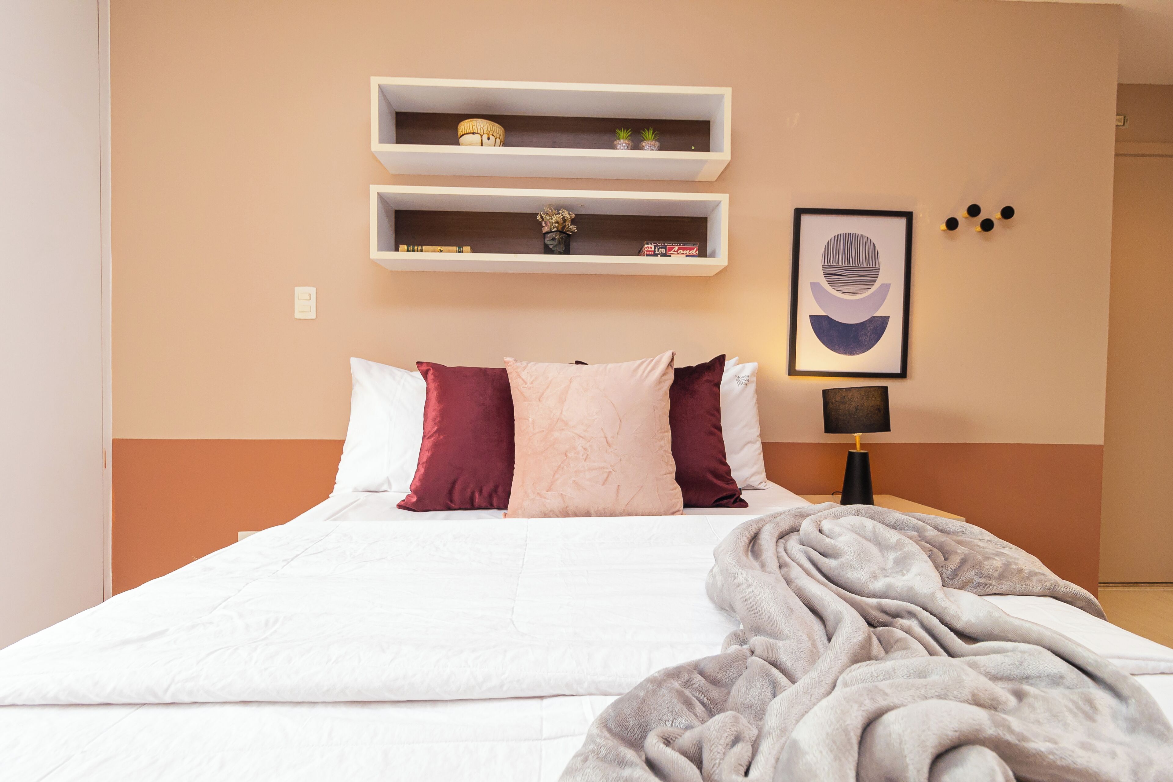 A neatly made bed with white and maroon pillows, a grey blanket, and two white shelves above it.