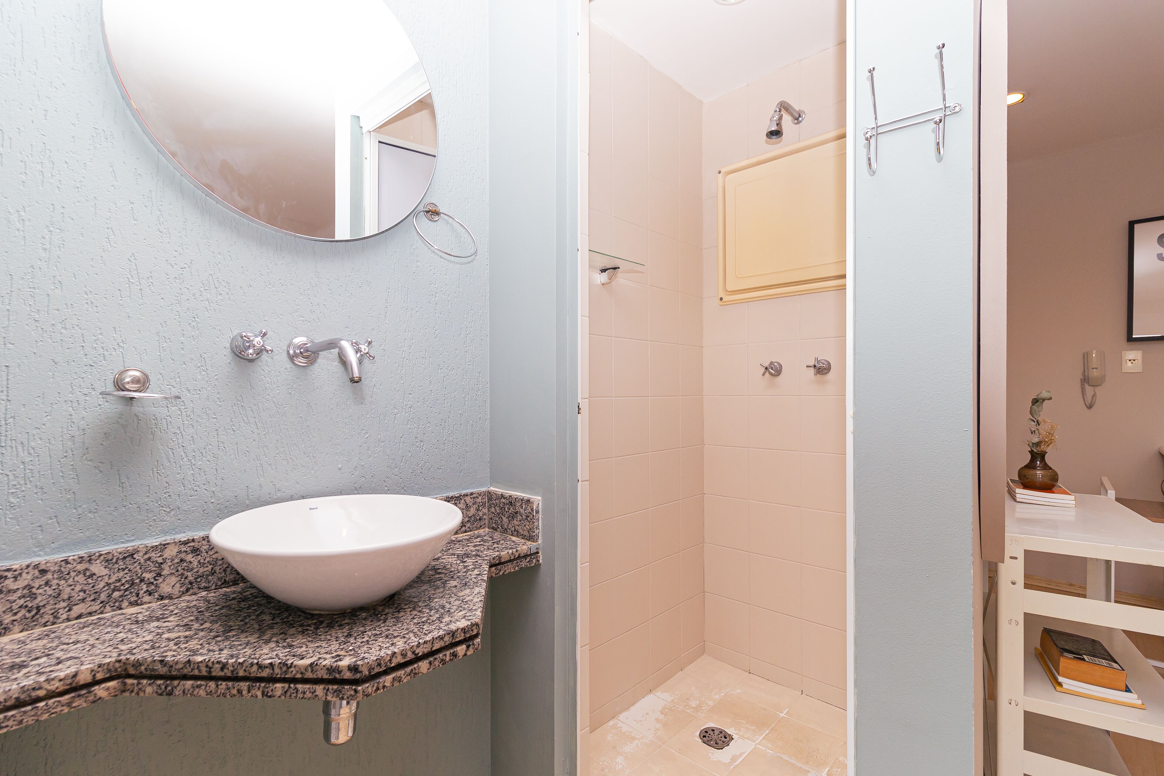 Une salle de bain avec un comptoir en granit, un lavabo blanc, un miroir, une douche avec une porte en verre et une étagère blanche contenant des livres.