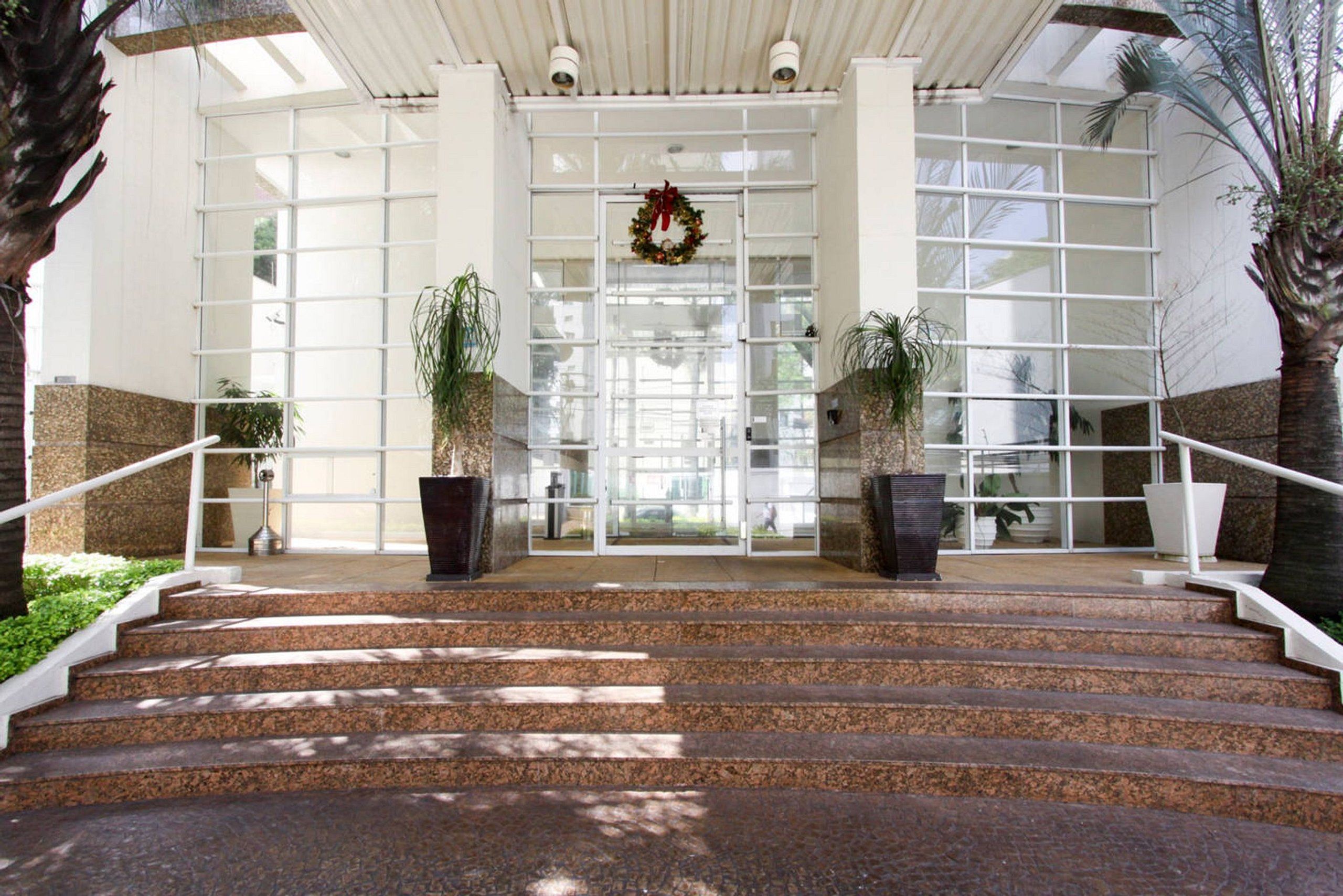 A hotel entrance with a set of stairs leading up to a glass door, flanked by two large potted plants and a wreath above the door.