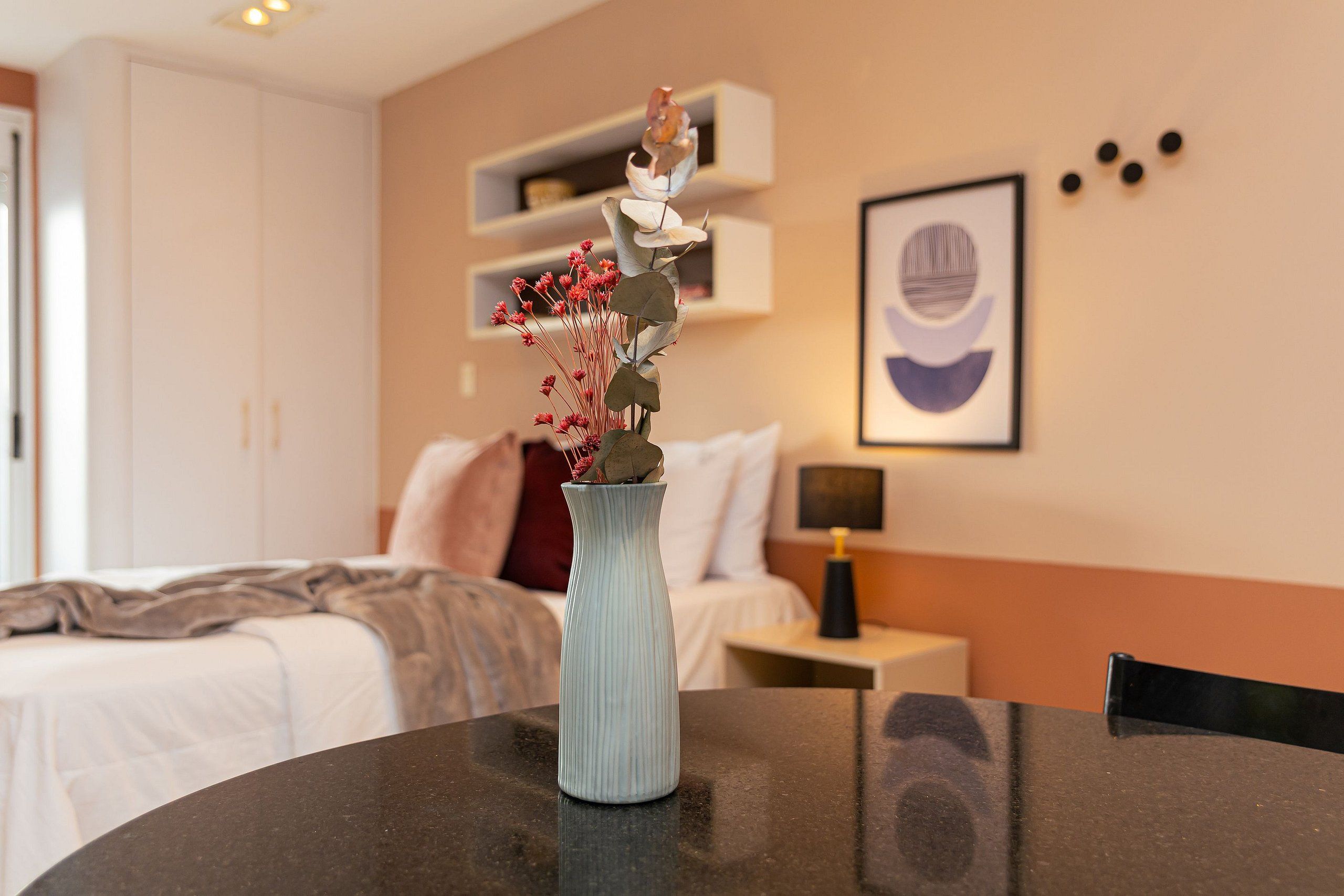 A hotel room with a bed, a nightstand, a lamp, a vase with flowers, and a wall with a framed picture.