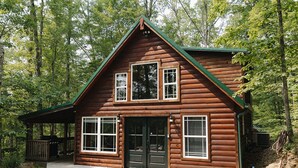 Une cabane en rondins à deux étages, dotée d’un porche, de grandes fenêtres et d’un toit végétalisé, entourée d’arbres.