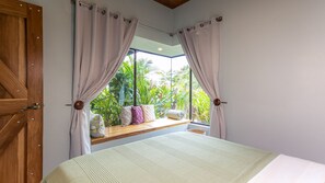 A bedroom with a large window, a wooden bench with cushions, and a bed with a white bedspread.