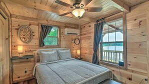 Un dormitorio de cabaña de madera con cama, ventilador de techo, ventana con cortinas y vistas al exterior.