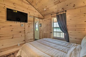 A wooden cabin bedroom with a bed, a mounted TV, a window with curtains, and a door.