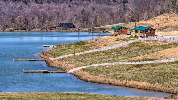 Una cabaña junto al lago con una terraza, rodeada de un paisaje natural y un bosque al fondo.