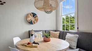 A round wooden table with a bottle of champagne, glasses, and a fruit platter. A gray sofa with patterned cushions is against a wall with a large window.