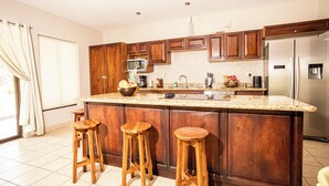 A kitchen with wooden cabinets, a stainless steel refrigerator, a granite countertop, and three wooden stools. - Condo Pacifico Quepos, 5 minutes from National Park Manuel Antonio (Quepos)