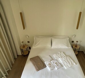 A neatly made bed with pillows, a folded robe, and towels on a white bedspread. There are two bedside tables with lamps and a small round table with a lamp.