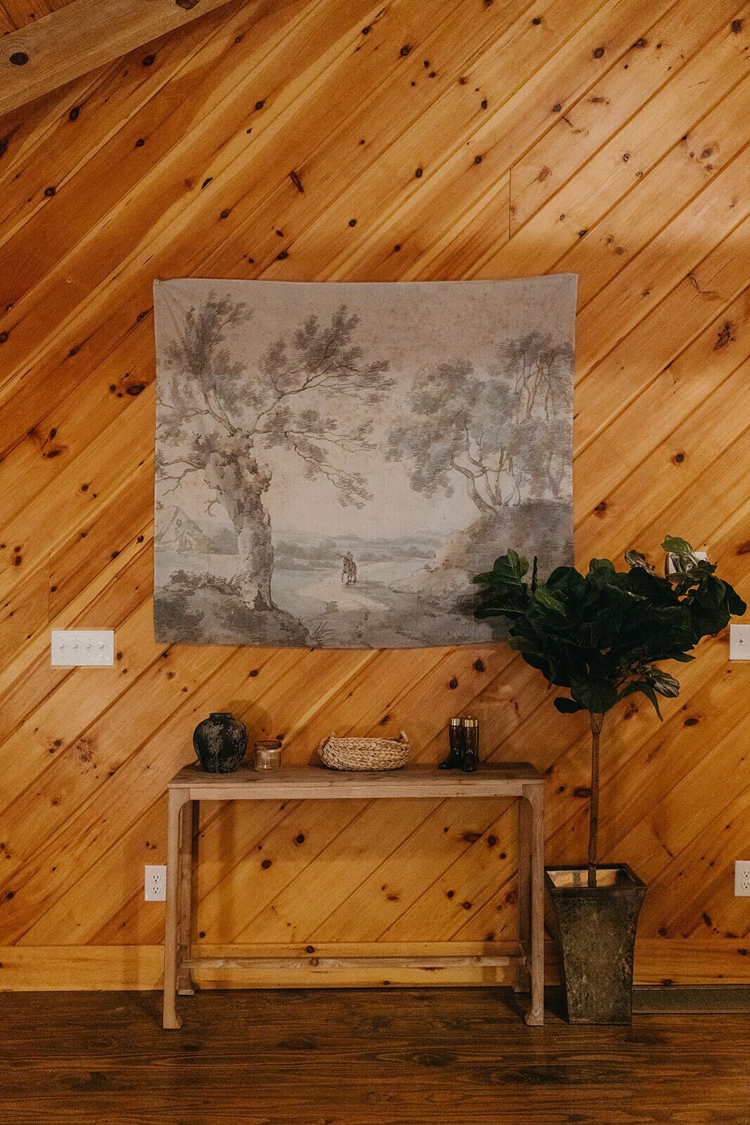 Pared de madera con un cuadro de un paisaje que muestra árboles y un sendero. Debajo del cuadro, una consola de madera con un jarrón, una vela y una planta pequeña.