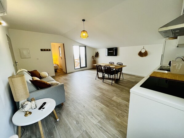 Una sala de estar moderna con un sofá gris, una mesita blanca con una lámpara, y un comedor con una mesa y sillas de madera.