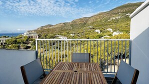 A wooden table and chairs on a balcony with a white railing, overlooking a hillside with buildings and greenery.