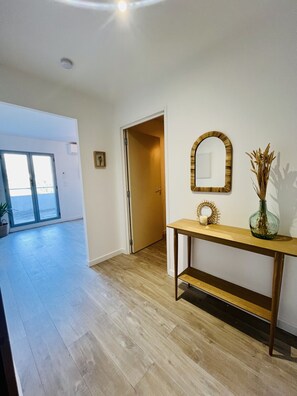 Una habitación amplia con piso de madera, una consola con espejo y objetos decorativos, una puerta que da a otra habitación y vistas a través de un ventanal.