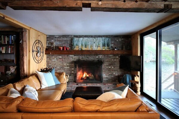 Cozy living room with a brick fireplace, leather sofa, and bookshelf, featuring a rustic ceiling and a large window with an outdoor view.