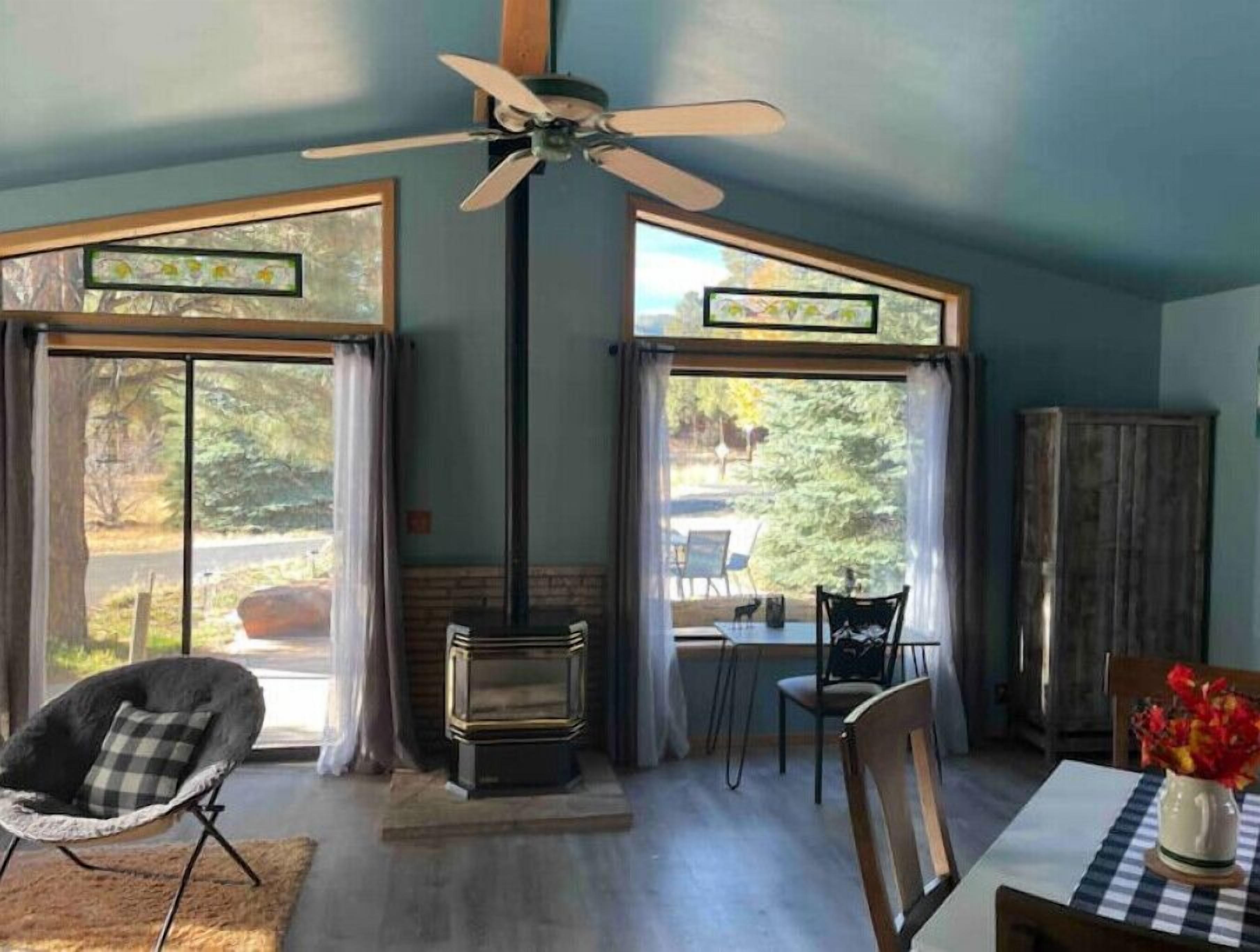 A room with a ceiling fan, large windows with curtains, a fireplace, and a dining table with chairs.