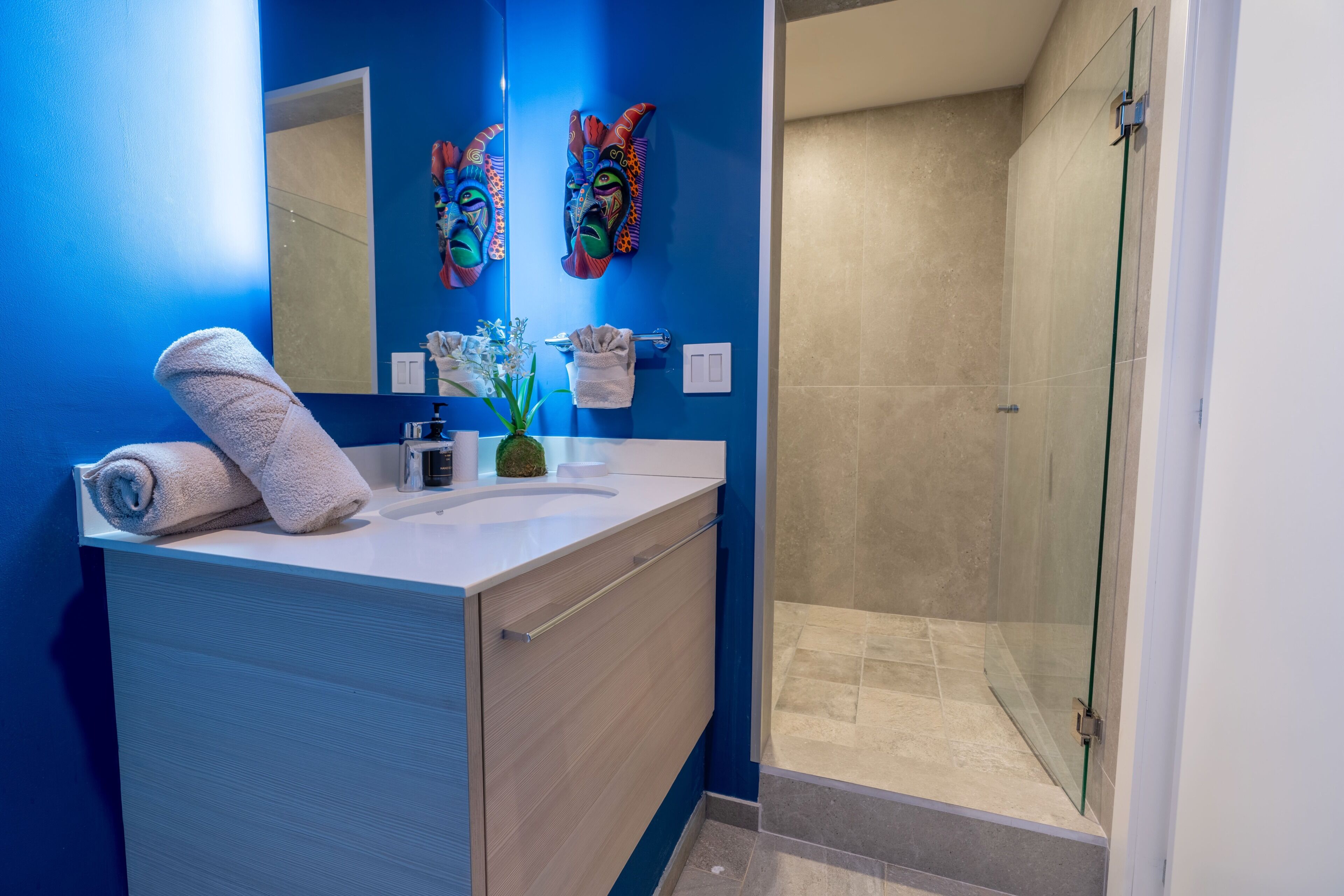 Un bany modern amb una paret blava, un lavabo blanc, un espai de banyera amb envidradora i màscares decoratives a la paret.