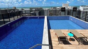 A rooftop pool with two separate sections, a wooden deck with lounge chairs and a small table, and a clear view of the ocean and buildings in the background. - Melhores Flats (João Pessoa)