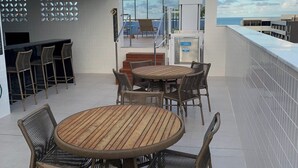 A rooftop terrace with a round wooden table and wicker chairs, a bar area with stools, and a view of the sea. - Melhores Flats (João Pessoa)