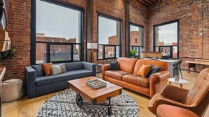 A spacious loft with exposed brick walls, large windows offering views of the city, a modern living area with sofas and a coffee table, and a dining area with a small table and chairs. - XCHNG 4 Apartments Downtown. Side by Side. (Effingham)