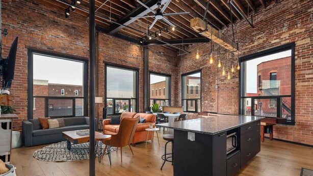 A spacious loft with exposed brick walls, large windows, a central kitchen island, and a cozy seating area. - XCHNG 4 Apartments Downtown. Side by Side. (Effingham)