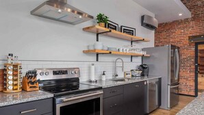 A modern kitchen with stainless steel appliances, wooden shelves, and a brick wall. - XCHNG 4 Apartments Downtown. Side by Side. (Effingham)