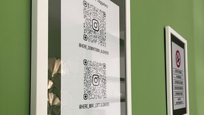 Un código QR enmarcado en una pared verde, con un cartel de prohibido fumar a su lado.