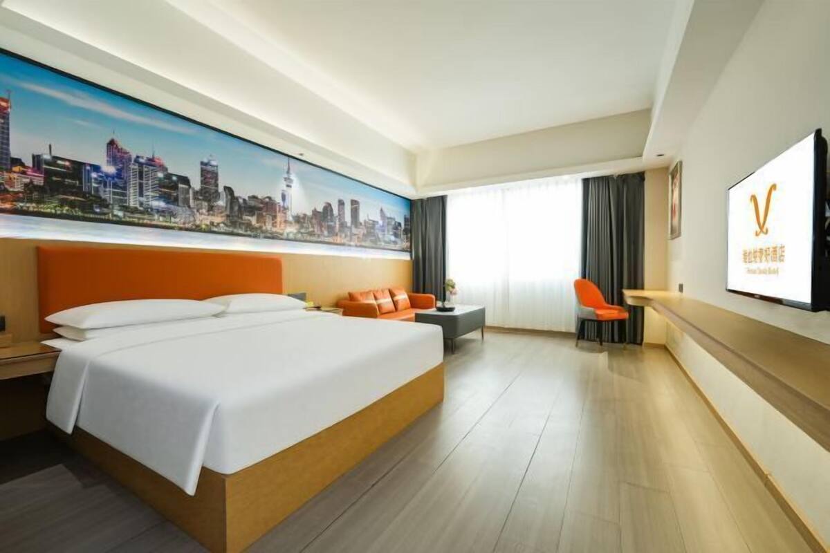 A hotel room with a large bed, a cityscape mural, a flat-screen TV, and a seating area with a chair and table.