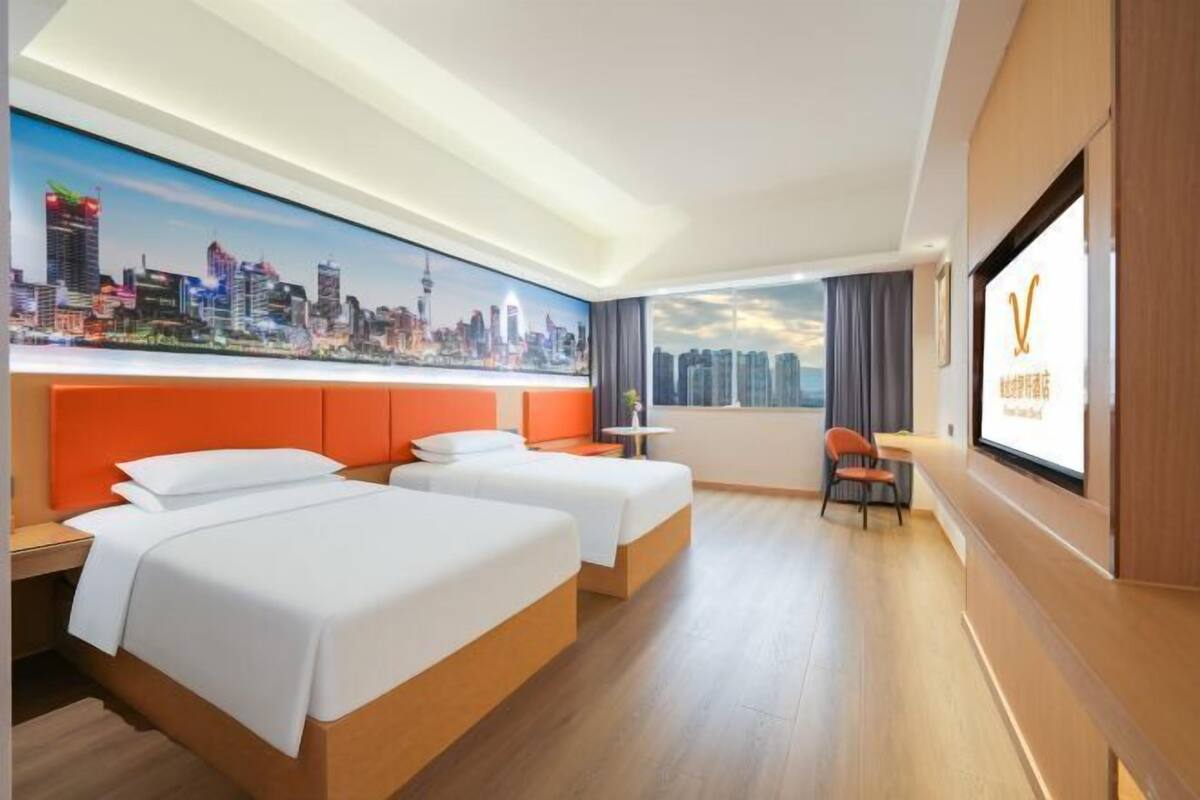 A hotel room with two beds, a cityscape mural, a large window with curtains, and a television screen.
