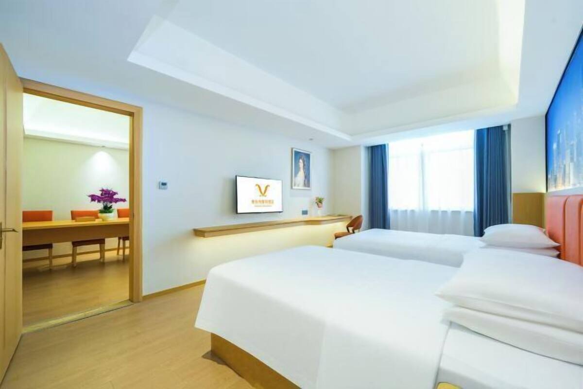 A hotel room with two beds, a desk with a chair, a small table with a vase, and a framed picture on the wall.