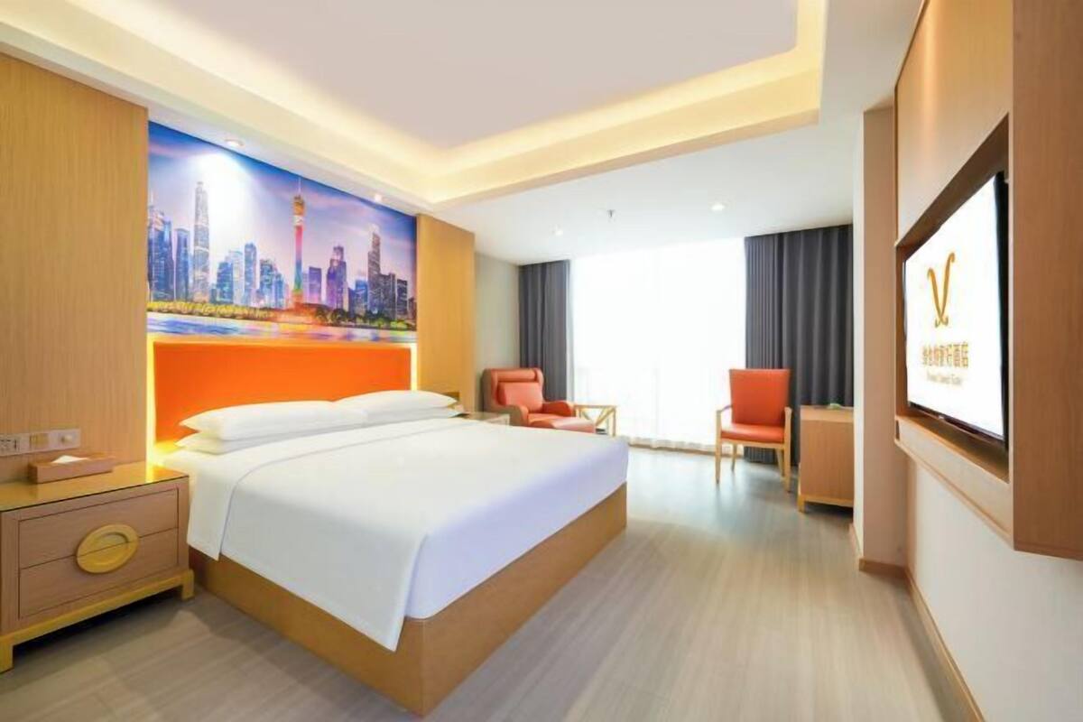 A hotel room with a bed, a desk, a chair, a TV, and a cityscape mural on the wall.