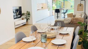 A dining area with a wooden table set for six, surrounded by chairs, with a TV and a kitchen in the background. - Modern townhouse - Private Garden (Differdange)