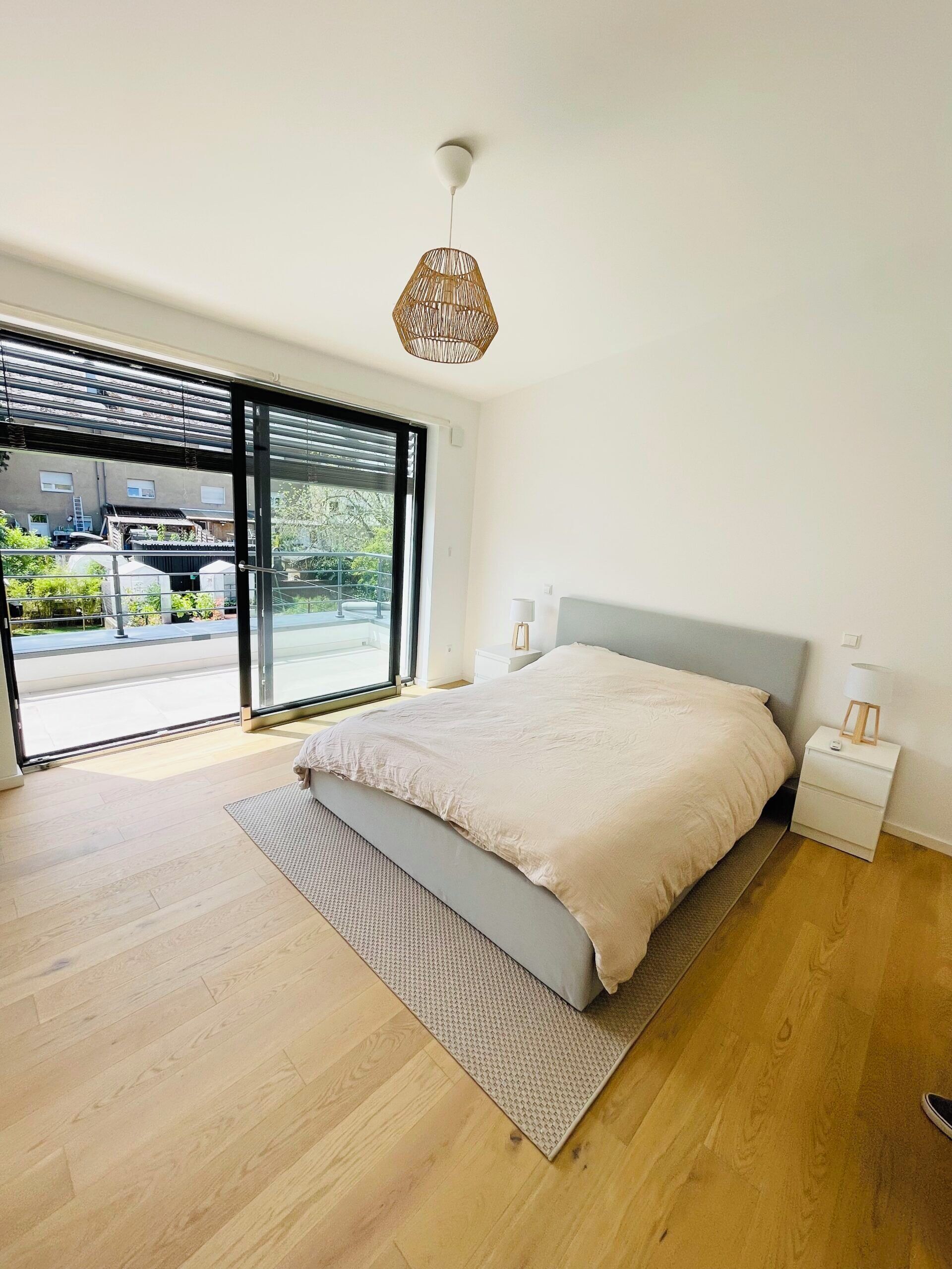 A modern bedroom with a large bed, a bedside table, a floor lamp, and a view through a sliding glass door to an outdoor area.