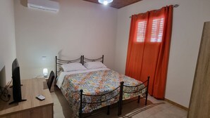 Een slaapkamer met een bed, een bureau met een computer, een stoel, een airconditioner en een raam met oranje gordijnen.