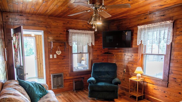 A cozy cabin interior with wooden walls, a ceiling fan, a television, and a fireplace.