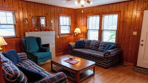A cozy living room with wooden walls, a fireplace, a teal armchair, a plaid sofa, and a coffee table.
