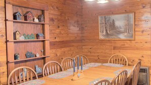 A wooden dining room with a table set for six, surrounded by wooden chairs. A wooden bookshelf with decorative items is to the left.