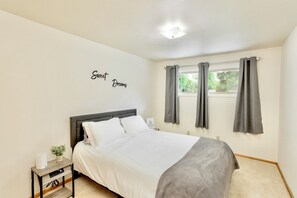 Un dormitorio con cama, mesita de noche, estante en la pared y ventana con cortinas.