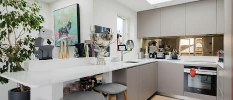 A modern kitchen with a white countertop, grey cabinets, and a skylight. There are bar stools, a large decorative bowl, and various kitchen appliances.