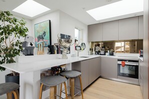 A modern kitchen with a white countertop, grey cabinets, and a skylight. There are bar stools, a large decorative bowl, and various kitchen appliances. - Spacious and Stylishly Designed 2BD Flat, Clapham (London)