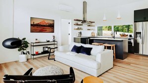 A modern living room with a white sofa, a coffee table, a black leather chair, and a black lamp. There is a kitchen area with a stove, sink, and cabinets. A large window provides a view of greenery outside.